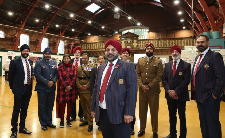 Man in turban smiles with members of the Armed Forces also dressed in turbans and members of the community in high-ceilinged building