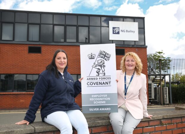 Two women holding a Silver Award banner outside the office of NG Bailey